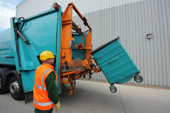 A man watches a refuse collection vehicle raise a large wheeled bin