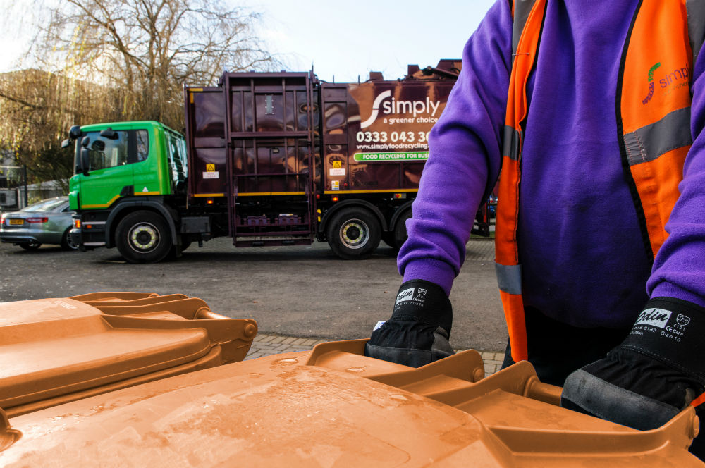 A man wearing a purple hoodie and orange hi-viz vest rests his hand on a brown wheelie bin, his head is out of shot, there is a foodcollection vehicle in the background